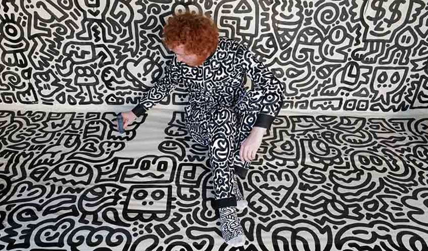 Artist Mr Doodle drawing with a pen while sat on the floor, with the floor and wall covered in black doodle art on a white background