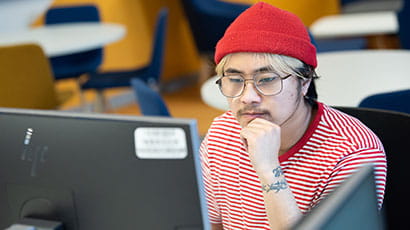 A student wearing a red beanie looks at a computer screen at a library workspace.