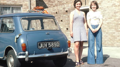 Two young women in 1970s fashions, standing next to a blue mini cooper car.
