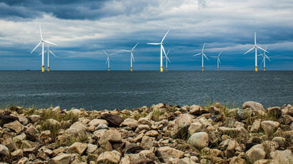 Wind turbines off the shore of the UK with a rocky beach in foreground.