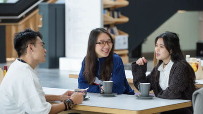 Three international students sitting at table in the Business school, having coffee and chatting to each other.