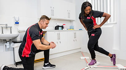 In a clinic treatment room, a rehabilitation specialist kneels in front of client, who is lunging with one leg outstretched to the side behind her.