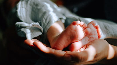 Feet of a newborn baby, held by the hand of an adult, with a shard of sunlight shining onto both the feet and the hand
