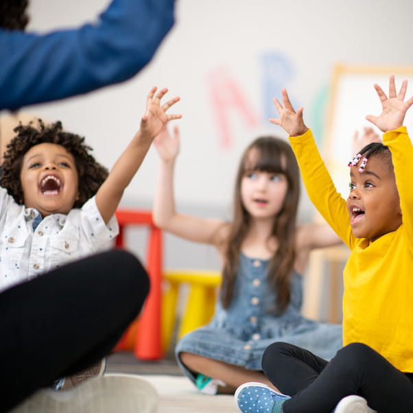 A multi-ethnic group of preschool students sitting with their legs crossed on the floor in their classroom with their teacher facing them. The happy kids are smiling and have their arms raised in the air.