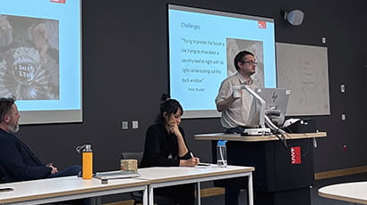 Professor James Green delivering his presentation at the RIPIL Inaugural Showcase Event on 6 December 2023. He is at the speaker's podium facing the audience with his presentation behind him. Suwita Hani Randhawa and Mike Pollard are seated.