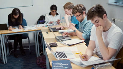 Group of students working at desks in a classroom, with laptops and notebooks