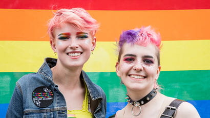 Two people standing in front of a rainbow flag at Bristol Pride.