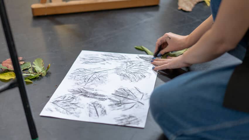 Student creating charcoal rubbings of leaves and natural materials on the floor
