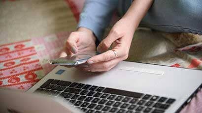 Close up of girl's hand holding mobile phone above a laptop whilst sat on the floor.