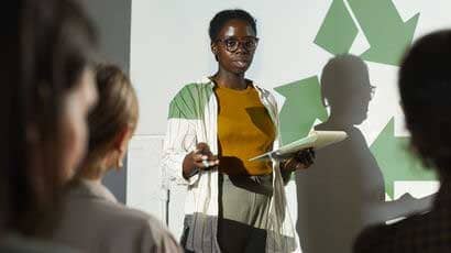 A young African-American woman presenting at the front of a lecture theatre with a presentation behind her.