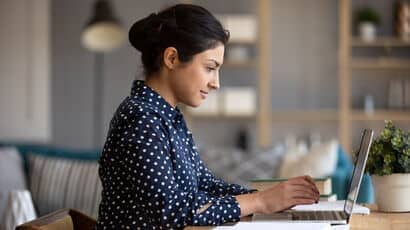 Young adult Indian woman in navy blue white polka-dotted long-sleeved top using laptop at a table.