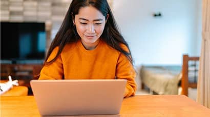 A young Chinese girl in an orange jumper sitting at a desk and looking at her laptop.