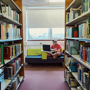 A student sat working at a laptop between rows of books in a library setting