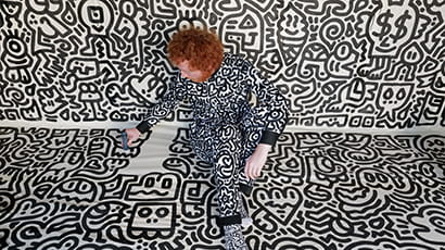 Mr Doodle, aka Sam Cox, wearing a suit covered in black and white squiggles, sitting in a room painted with the same pattern on the walls and floor.