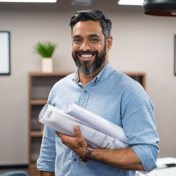 Man in blue shirt, standing to camera in office holding plan drawings under arm. 