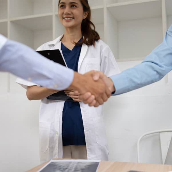 A woman in white coat standing behind two arms shaking hands.