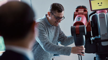 Man wearing glasses demonstrates his robotics project while a man in a suit looks on.