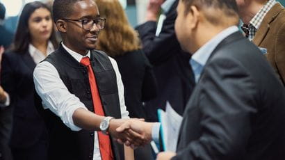 A student and visitor shaking hands at the Dragons' Den event, with a small crowd in the background.