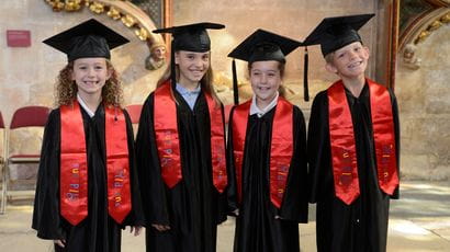 Four primary school aged children standing in a row, dressed in graduation gowns and wearing mortar boards.