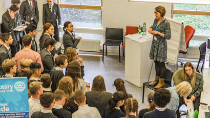 Female staff member standing on chair, talking to a large group of school students.