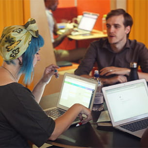 Three people meeting around a table with laptops engaging in a discussion.
