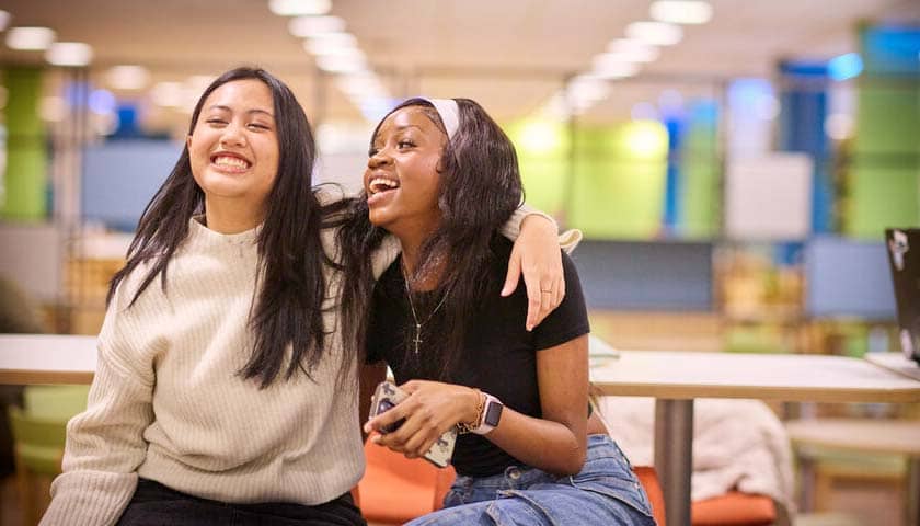 A couple of smiling students sat on a desk one with their arm around the other. 