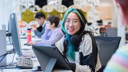 Girl with headset around her neck sat at a pc smiling at a person adjacent.