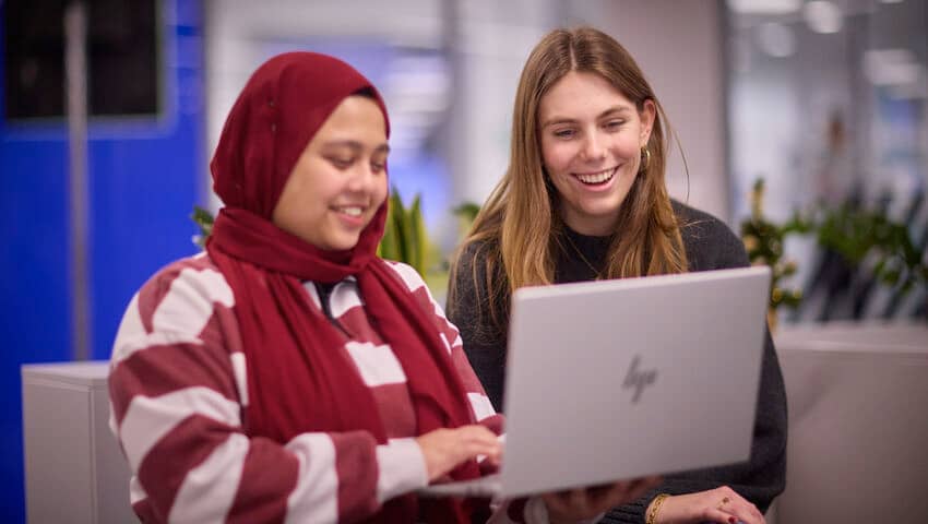 Couple of students sat on a sofa smiling whilst looking at a laptop