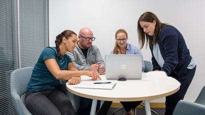 Group of three people sitting, and one standing, around a table, viewing a laptop.