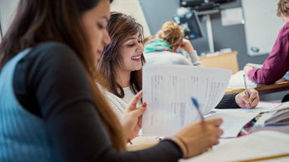 Two students in a classroom, writing on worksheets and smiling