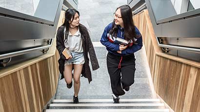 Two international students walking up some stairs in the Business School, chatting to each other.