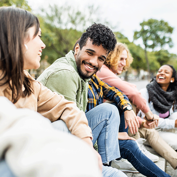 A group of four students, sat on a set of steps outside, laughing with trees in the background.