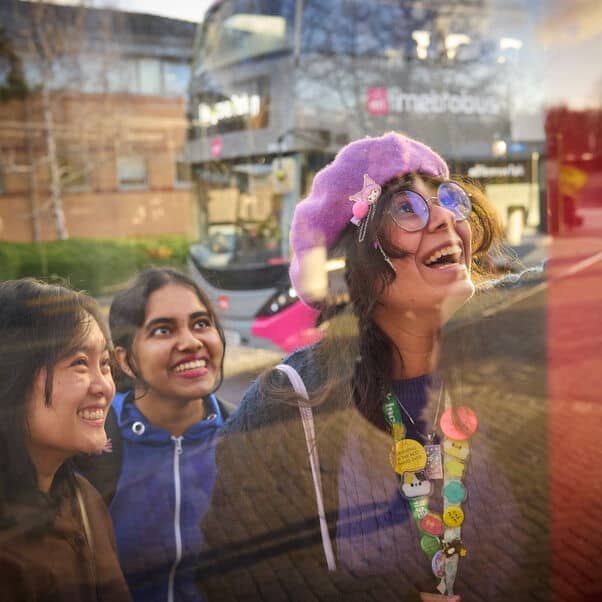 three young women looking at the bus timetable, smiling; a bus going past in the background