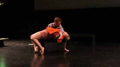 Actors on a bare stage, one swooning, one holding the other