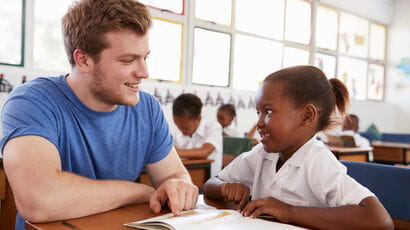 Teacher and primary school student sitting at a classroom table discussing a book.