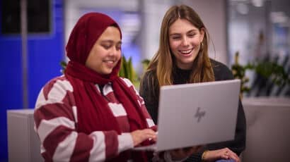 Two females, one Caucasian and one Muslim wearing a head scarf, sitting next to each other and looking at a laptop.