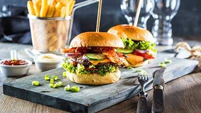 Burgers on a serving board with chips, dips and glasses in the background.