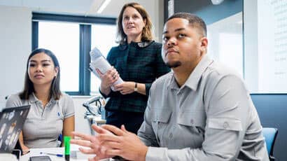 Students sat in a class with a lecturer standing behind them holding papers.