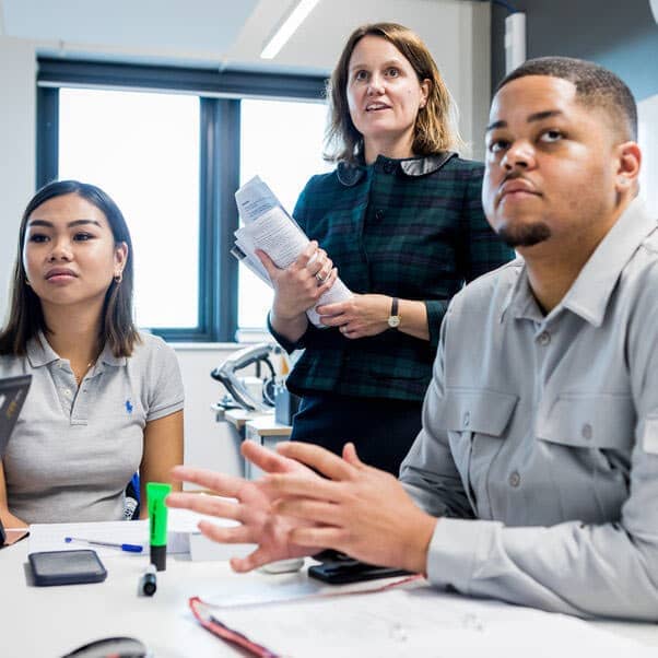 A couple of students sat at a desk looking to the front with an academic standing behind them.