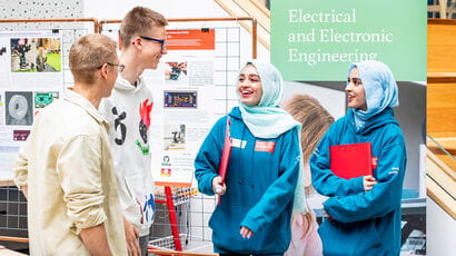 Two student ambassadors chat to a prospective student and their family at an Open Day in front of a banner that says 'electrical and electronic engineering'.