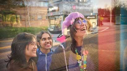 Three students looking at the bus timetable, smiling; a bus going past in the background