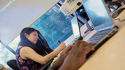Close up of a persons hands working on a laptop with another person working on a laptop in the background
