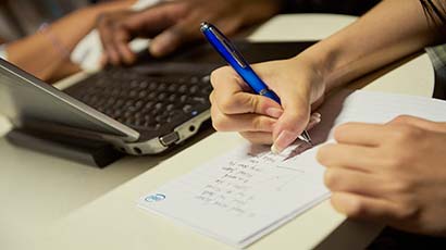 Close up of a student writing on a notepad and another student using a laptop