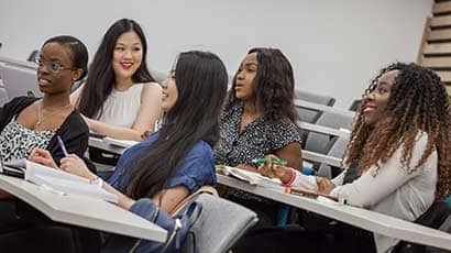Group of students in a lecture, with books, pens and laptops, all are wearing smiles and interacting with the off screen lecturer.