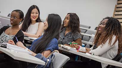 Group of students in a lecture, with books, pens and laptops, all are wearing smiles and interacting with the off screen lecturer.