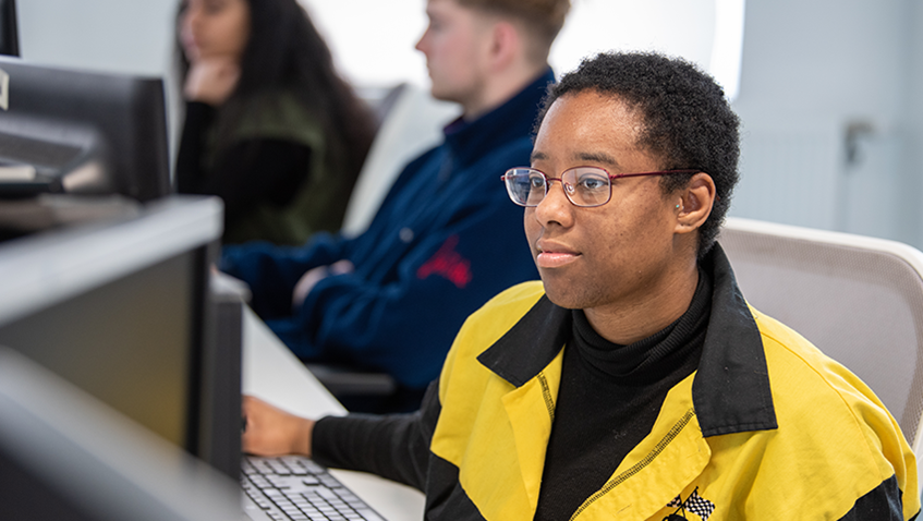 Student in black and yellow jacket watching something on a computer screen on campus.