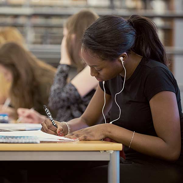 A student at a desk with headphones in, writing in a notepad.