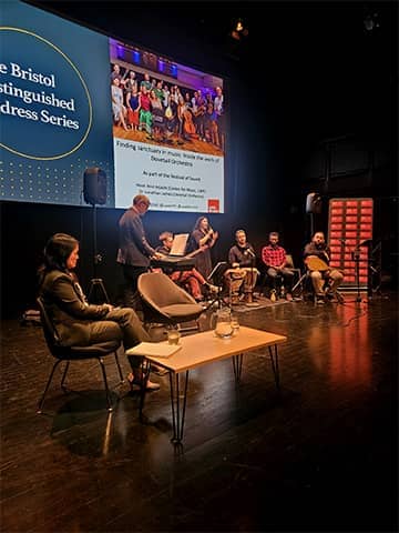 Panel discussion on a theatre stage with seated speakers, musicians, and instruments, with a large screen behind displaying “The Bristol Distinguished Address Series”.