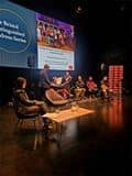 Panel discussion on a theatre stage with seated speakers, musicians, and instruments, with a large screen behind displaying “The Bristol Distinguished Address Series”.