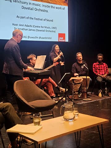 A performer singing into a microphone on stage, accompanied by musicians seated nearby.
